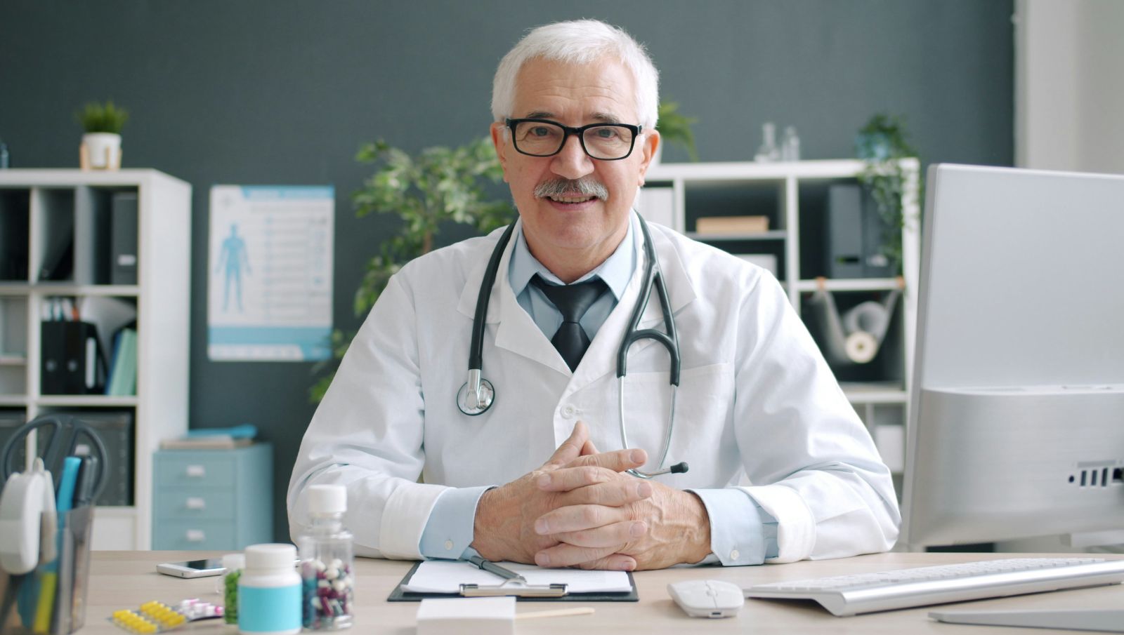 A smiling, senior male doctor in a white lab coat sitting at a wooden desk in a bright office, representing professional medical cannabis consultations in Daphne, Alabama.