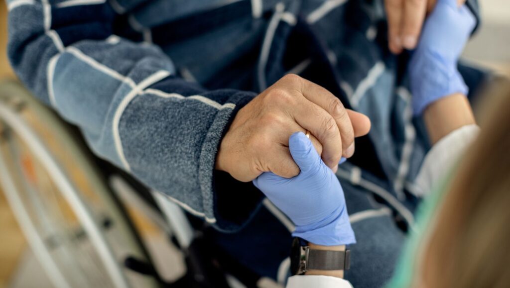 An elderly man in a wheelchair holding the hand of his caregiver, representing medical cannabis support for Alabama residents with ALS, MS, and spinal cord injuries.