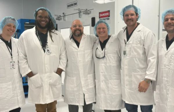 Homestead Health staff members wearing cleanroom suits, hairnets, and masks, smiling while standing together in a sterile facility.